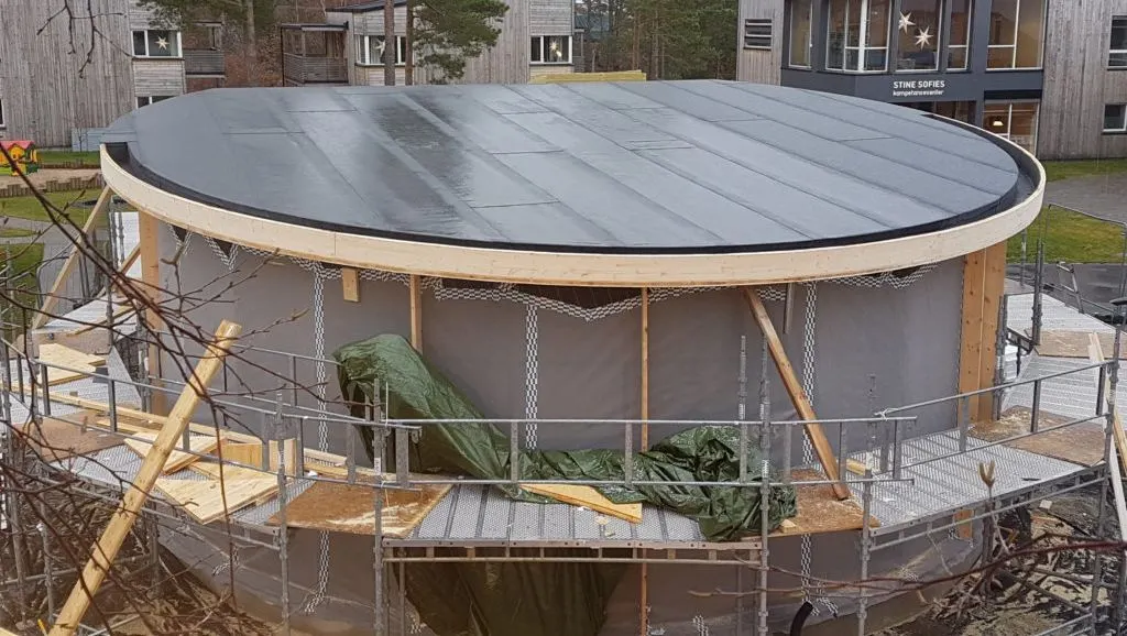 Rund bygning under oppføring med ferdig montert takbelegg og stillas rundt.