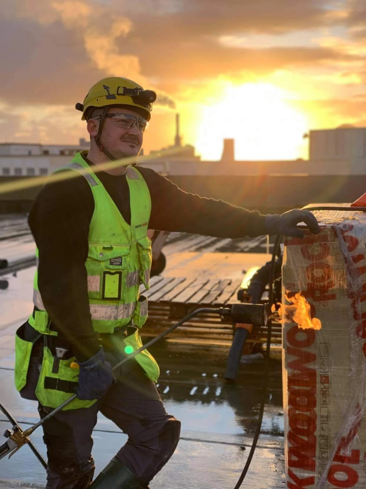 Takdekker i verneutstyr og hjelm på byggeplass i solnedgang.